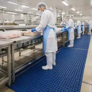 higienske-podloge-za-zivilsko-industrijo-slika-1 Delavec v živilskem obratu stoji na modri protizdrsni podlogi ob proizvodni liniji, kjer higienske podloge za živilsko industrijo pomagajo zagotavljati čistočo, varnost in udobje pri delu.