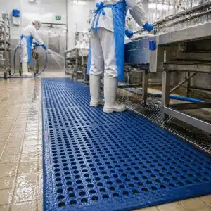 higienske-podloge-za-zivilsko-industrijo-slika-2 Mokra delovna cona v živilskem obratu z modro odtočno talno oblogo, kjer higienske podloge za živilsko industrijo izboljšujejo varnost, odvajanje tekočin in udobje pri delu.